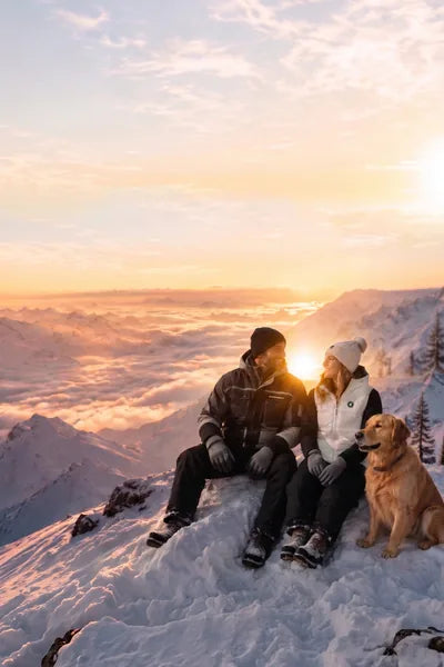 Paar mit Hund auf einem verschneiten Berggipfel bei Sonnenuntergang in winterlicher Landschaft